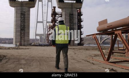Man in uniform and hardhat walking on dusty path under unfinished bridge during work on construction site Stock Photo