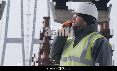 Man in hardhat and goggles inspecting unfinished bridge and talking on smartphone with boss during work on construction site Stock Photo