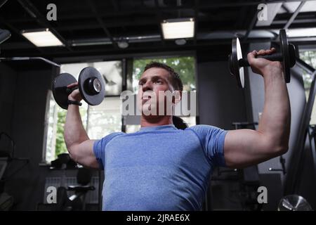 Focused muscular young man lifting dumbbells, doing shoulders exercise Stock Photo