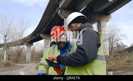 Mature and adult man using tablet and talking while standing near concrete bridge in outskirts Stock Photo