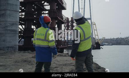 Men in uniform inspecting unfinished bridge over river while working on construction site Stock Photo