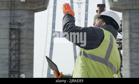 Men in uniform browsing data on tablet and discussing unfinished bridge during work on construction site Stock Photo