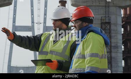Men in uniform browsing data on tablet and discussing unfinished bridge during work on construction site Stock Photo