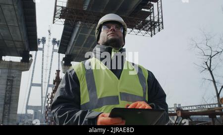 Male builder browsing data on tablet while standing under unfinished bridge during work on construction site Stock Photo
