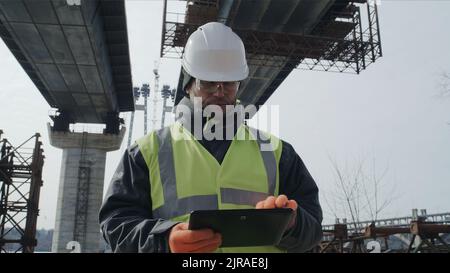 Male builder browsing data on tablet while standing under unfinished bridge during work on construction site Stock Photo