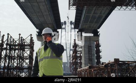 Man in uniform and hardhat answering phone call while walking under concrete bridge during inspection on construction site Stock Photo