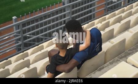 From above African American man embracing mixed race boy and speaking while sitting on plastic seats and watching match together on stadium Stock Photo