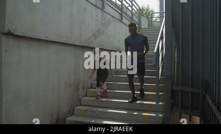 Pan left of black man and mixed race boy running on staircase during workout on stadium in summer Stock Photo