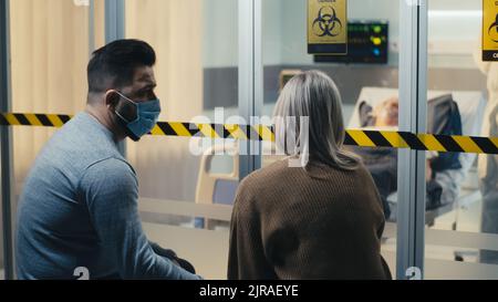 Man in mask supporting crying woman while sitting near glass wall of coronavirus ward while doctor in hazmat suit taking care of elderly patient Stock Photo