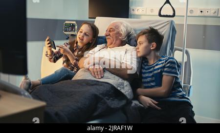 Aged man smiling and talking with boy and girl while lying on bed and watching video on mobile phone in ward of clinic Stock Photo