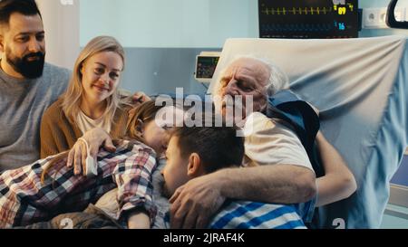 Aged male patient embracing kids and talking with adult man and woman while lying on bed in ward of modern hospital Stock Photo