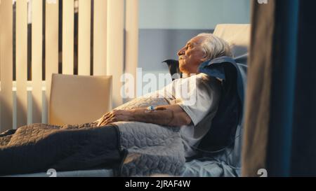 Pan left view of lonely elderly man lying on bed and looking away in ward of modern clinic Stock Photo