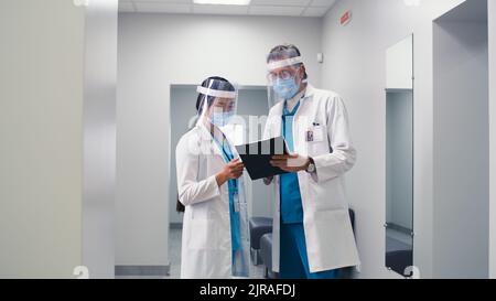 Diverse man and woman in medical uniform and face shields discussing data on clipboard then walking different direction in hallway of contemporary hospital Stock Photo
