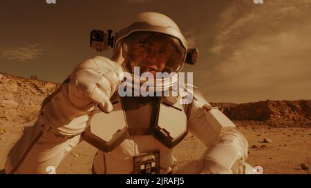 Man in spacesuit walking and turning on camera then waving hand and pointing away while speaking and shooting video during colonization of Mars Stock Photo