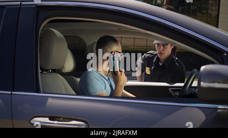 Angry man finishing phone call then talking and showing driver license to serious woman in police uniform while sitting in car on city street Stock Photo