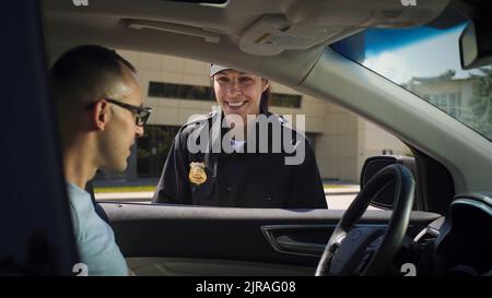 Cheerful woman in police uniform friendly smiling and checking documents of man sitting in car on city street Stock Photo