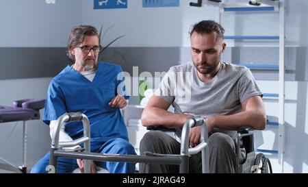 Middle aged rehabilitation therapist talking with depressed man on wheelchair in hospital Stock Photo