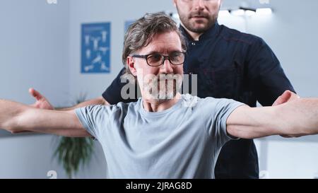 Man supporting middle aged male patient doing exercise with dumbbells during rehabilitation session in clinic Stock Photo