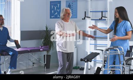 Adult woman helping senior male patient to stroll without walking frame while mature colleague using tablet then doctors clapping hands during rehabilitation session in hospital Stock Photo
