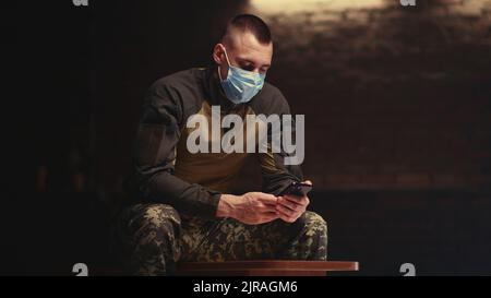 Military man in mask waving hand and speaking while sitting on bench and making video call on smartphone in dark room Stock Photo