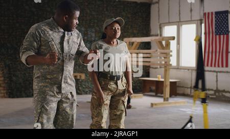 African American military woman doing exercise with training ropes and listening to male instructor in camouflage during suspension workout in gym of USA military base Stock Photo