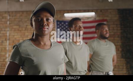 African American female soldier turning head and looking at camera while standing near male squadmates against brick wall with USA flag on military base Stock Photo