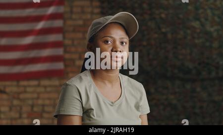 Black female soldier in t shirt and cap looking at camera while resting against brick wall with USA flag on military base Stock Photo