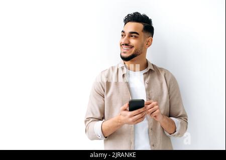 Confident positive handsome indian or arabian young man, holding smartphone in hand, chatting online, browsing internet, looking happily to the side, standing on isolated white background, smiling Stock Photo