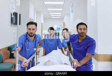 doctors and nurses wheeling elderly patient in gurney on hospital corridor at emergency department. Work of emergency medical team and paramedics Stock Photo