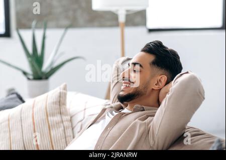 Close-up of satisfied happy calm indian or arabian young man, resting in the living room on the sofa, holds his hands behind his head with closed eyes and smiles. Chilling and relaxing at home. Stock Photo