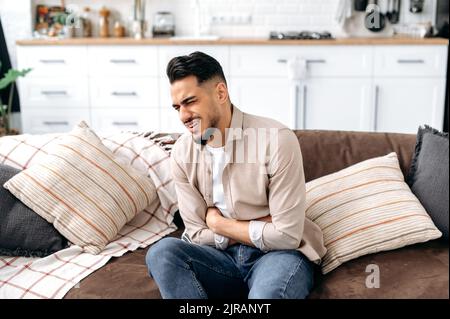 Frustrated unhealthy indian or arabian young man, sitting on the sofa in the living room, holding his stomach, suffering from spasm, stomach pain, poisoning, needs treatment, sad facial expression Stock Photo