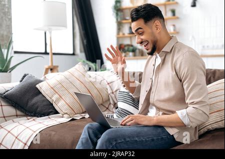 Cheerful positive arabian or indian young stylish modern man, spending time at home, sitting on sofa in living room, talking on video call with friends, family or colleague, waves hand, smiling Stock Photo