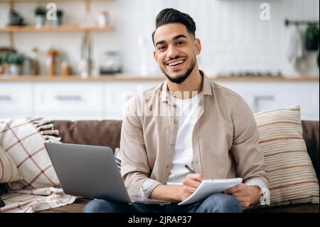 Handsome friendly indian or arabian man taking notes while working on project or learning online, listening webinar uses laptop, sitting on a sofa in a living room, prepares a report, looks at camera Stock Photo