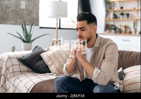 Worried confused upset indian or arabian young man in casual wear, sitting alone on the sofa at home in the living room, experiencing personal problems, worries, depression, stress Stock Photo