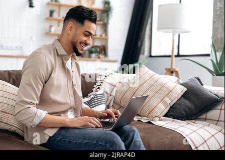 Glad positive arabian or indian young stylish modern man, spending time at home, sitting on sofa in living room, using laptop, browsing internet, social media, watching videos, smiling Stock Photo