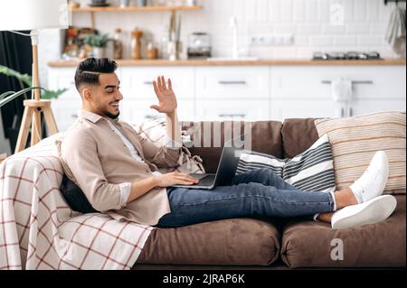 Happy indian or arabian young man, freelancer or student, sits on comfortable sofa at home in living room, talking on video call with friend, colleague, client, smiling .Online video communication Stock Photo