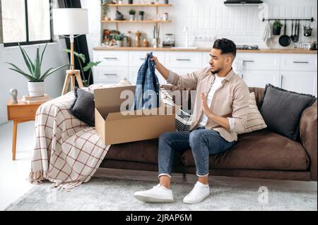 Disappointment arabian or indian stylish man, sitting on the sofa in the living room, unpacking his parcel, taking out clothes from the cardboard box, looking at her in frustration, dissatisfied Stock Photo