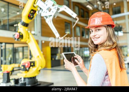 Smiling young female worker controlling robot arm with digital tablet Stock Photo