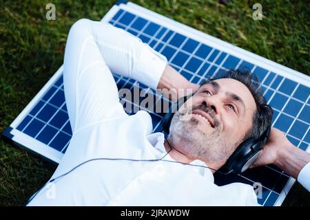 Smiling businessman with hands behind head lying on solar panel listening music through headphones Stock Photo