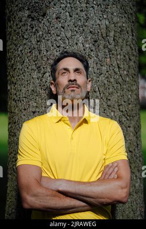 Thoughtful man with arms crossed leaning on tree trunk Stock Photo