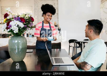 Smiling waitress serving coffee to customer sitting with laptop in cafe Stock Photo