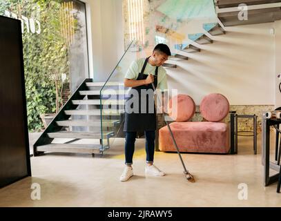 Young man wearing apron cleaning floor with broom in cafe Stock Photo