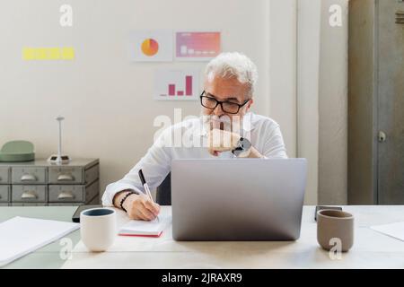Senior businessman with hand on chin writing in notepad at office Stock Photo