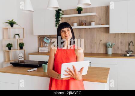 Thoughtful woman with tablet PC standing in kitchen at home Stock Photo