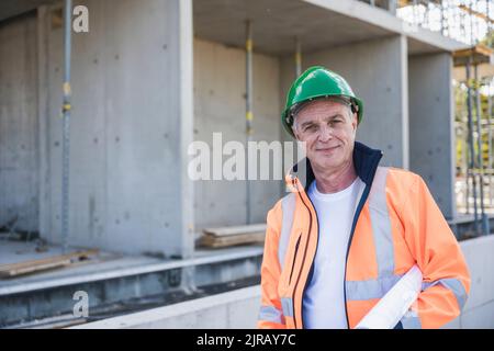 Smiling building contractor with blueprint standing at construction site Stock Photo