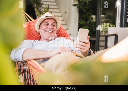Happy senior man using phone lying in hammock Stock Photo