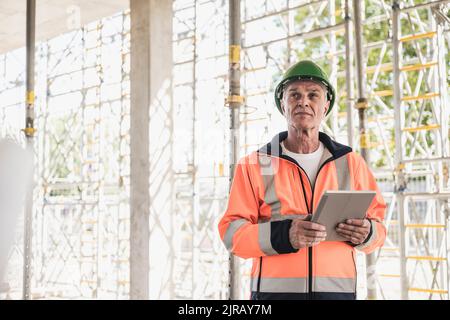 Thoughtful contractor holding tablet PC standing in front of scaffolding Stock Photo