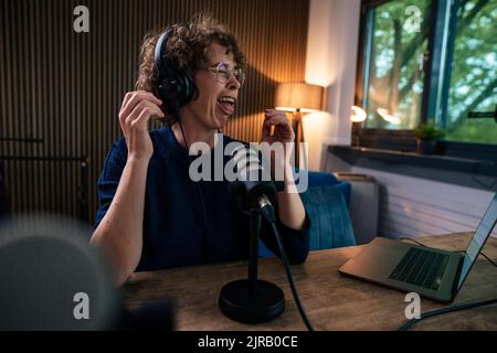 Presenter wearing headset making face sitting with microphone and laptop at desk in recording studio Stock Photo