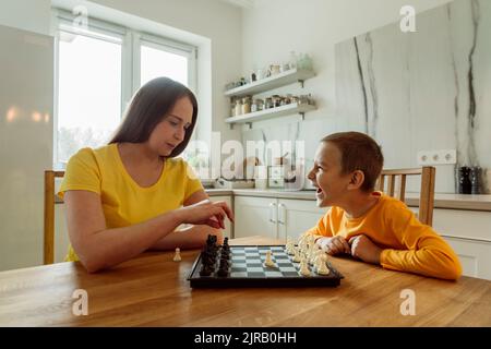 Happy son looking at mother playing chess in kitchen Stock Photo