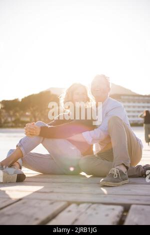 Cheerful mature couple sitting at jetty on sunny day Stock Photo